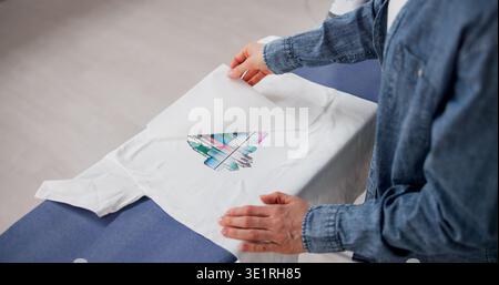 Femme appliquant la conception personnalisée de transfert de chaleur sur T-shirt avec fer à repasser à la maison Banque D'Images