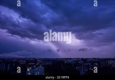 Paysage urbain nocturne spectaculaire sous un ciel turbulent et orageux. Nuages sombres illuminés par des éclairs, projetant une lueur violette étrange sur la scène. Les lumières de la ville des bâtiments et des rues créent du contraste. Banque D'Images