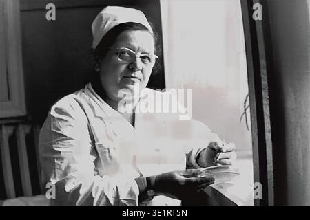 Une photographie d'archives en noir et blanc prise en 1977 montre une femme professionnelle de la santé portant des lunettes, un manteau blanc et une casquette, travaillant soigneusement près d'une fenêtre lumineuse à l'hôpital-usine AIZ à Slavyansk. L’infirmière ou le technicien de laboratoire spécialisé tient une boîte de Pétri et un petit bâton, effectuant un test de laboratoire ou une analyse bactériologique. Ce portrait intime souligne le dévouement et la précision requis dans les soins de santé soviétiques. Cet instantané historique documente la pratique clinique quotidienne, la médecine du travail et le Donbass pacifique avant la guerre. Banque D'Images
