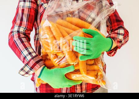 Agriculteur tenant un grand sac de carottes fraîches, portant des gants de travail verts et chemise à carreaux, concept de récolte agricole, focus sélectif Banque D'Images