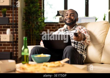 Afro-américain freelance travaille à distance à partir d'un canapé avec un appareil numérique sur ses genoux, en utilisant la télécommande pour régler le volume de la télévision. Jeune homme multitâche entre ordinateur portable et regarder la télévision à la maison. Banque D'Images