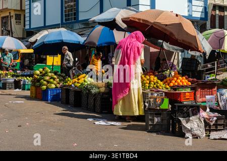 Scène de marché de rue animée à Udaipur avec des vendeurs vendant des fruits tropicaux sous des parapluies colorés près d'un bâtiment bleu et blanc. Banque D'Images