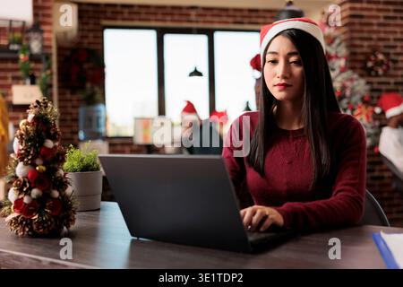 Chef d'entreprise de femme asiatique portant chapeau de père noël tapant sur ordinateur portable dans l'espace de travail décoré de noël. Employé assis au bureau et travaillant sur ordinateur au bureau de fête du nouvel an Banque D'Images