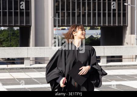 Diplômée asiatique debout sur la place carrelée portant une casquette de maintien de robe noire et un smartphone rose. Toit, extérieur, urbain, terrasse, vent soufflé, célébration, réussite || autorisation du modèle Banque D'Images