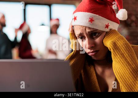 Triste femme caucasienne malheureuse portant le chapeau de santa travaillant sur ordinateur portable dans le bureau à la veille de vacances de noël. Employé épuisé de l'entreprise résolvant la tâche sur l'ordinateur pendant la saison de la nouvelle année Banque D'Images