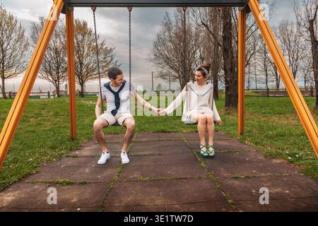 Jeune couple assis sur des balançoires se tenant la main, profitant de la vie commune dans un parc Banque D'Images