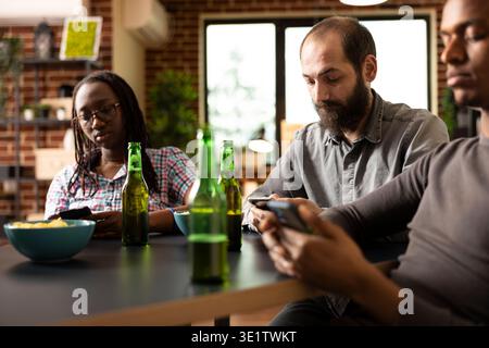 Individu masculin caucasien utilise un téléphone portable pour naviguer en ligne, avec une bouteille de bière placée sur la table devant lui. Homme blanc barbu avec appareil mobile, est assis au milieu d'amis, profitant de leur compagnie. Banque D'Images