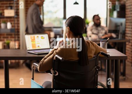 Vue arrière d'une employée en fauteuil roulant, utilisant son ordinateur portable et parlant avec un client de garde. Femme d'affaires handicapée avec un téléphone fixe à son oreille, travaillant sur une présentation de projet. Banque D'Images