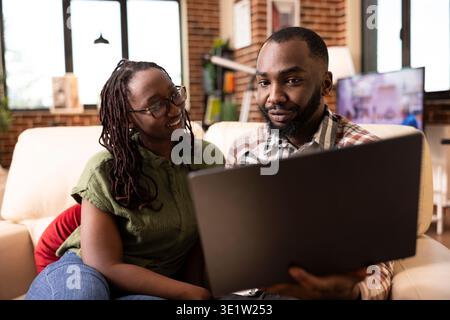 Petit ami et petite amie assis sur le canapé examinant l'itinéraire de voyage et les notes de travail sur ordinateur portable. Conjoints afro-américains multitâches, mélangeant la planification personnelle et le travail à distance dans un appartement confortable de mur de briques Banque D'Images