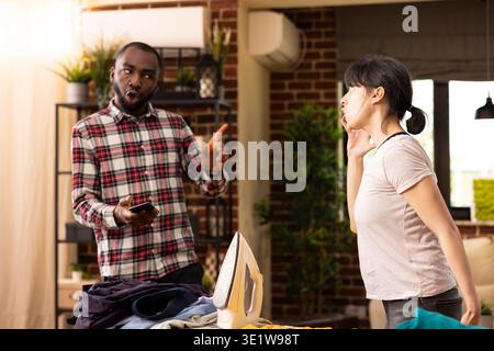 Couple marié diversifié ayant une discussion chauffée le jour de la lessive dans un appartement moderne. La petite amie blanche frustrée et le petit ami noir bouleversé expriment leur désaccord sur le partage des tâches ménagères à l'intérieur. Banque D'Images