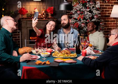 Happy People prend des photos ensemble à la maison, profitant d'un festin de dîner avec des verres d'alcool entouré de décorations de Noël. Divers amis et famille prenant des photos pendant la célébration de noël. Banque D'Images