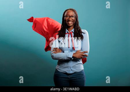 Plein d'assurance puissant jeune adulte porte un costume de super-héros sur fond bleu. Portrait d'une femme courageuse heureuse avec des superpuissances portant une cape héro rouge tout en se tenant debout avec les bras croisés. Banque D'Images