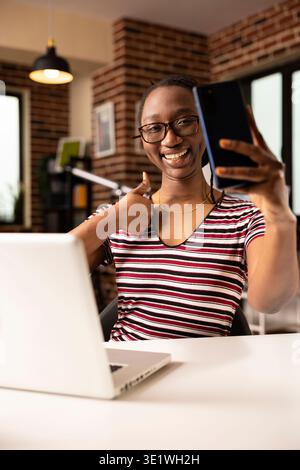 Joyeuse femme afro-américaine tenant smartphone, posant avec le pouce vers le haut geste tout en capturant selfie. Entrepreneure joyeuse assise au bureau, mettant à jour les médias sociaux avec une photo amusante à la maison. Banque D'Images