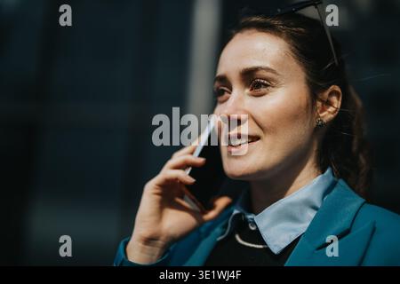 Femme d'affaires confiante sur un appel mobile, souriant à l'extérieur dans un blazer sarcelle et chemise bleu clair Banque D'Images