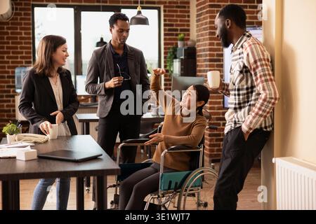 Employés professionnels ayant une conversation allégée, favorisant le travail d'équipe dans un contexte d'entreprise. Femme asiatique joyeuse dans un fauteuil roulant discutant avec son équipe inclusive, prenant une courte pause. Banque D'Images