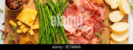 Une planche de charcuterie magnifiquement agencée avec des tranches de jambon, des blocs de fromage, des asperges fraîches et des noix croquantes. Parfait pour déguster avec frien Banque D'Images