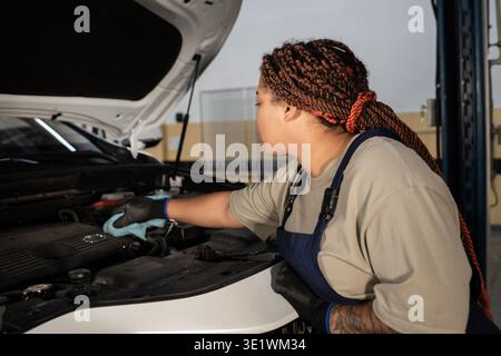 Jeune femme qualifiée ajuste les composants du moteur tout en travaillant dans un garage de réparation automobile. Banque D'Images