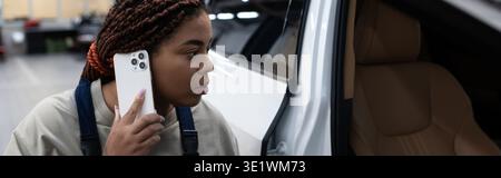 Jeune femme afro-américaine concentrée sur la réparation d'un véhicule au téléphone, bannière Banque D'Images