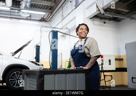 Une jeune femme habile en salopette sourit tout en manipulant des outils dans un atelier de réparation automobile occupé. Banque D'Images