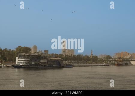 Vue sur le fleuve du Nil au Caire, en Égypte, avec un bateau de croisière à plusieurs ponts amarré le long du front de mer et une toile de fond de bâtiments modernes sous un ciel clair Banque D'Images