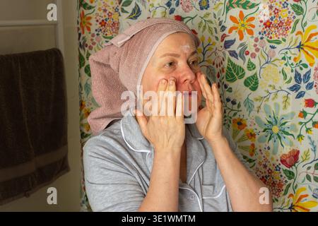 Une femme de 50 ans prépare sa peau en appliquant de la crème dans sa salle de bain. Elle porte une serviette sur la tête et se tient devant un curtai de douche coloré Banque D'Images