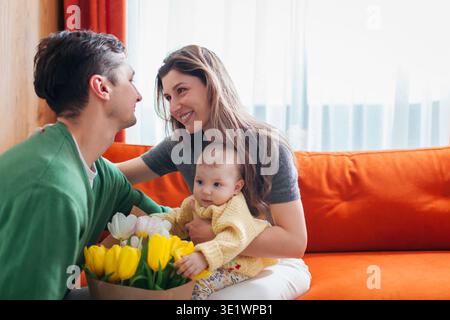 Bonne fête des mères. Mari surprenant sa femme avec un bouquet de fleurs à la maison. Femme tenant bébé fille en bas âge. Parents aimants Banque D'Images