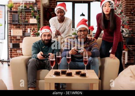 Heureux coworkers groupe célébrant noël ensemble en jouant au jeu vidéo dans un bureau décoré festif. Collègues avec manettes de jeu s'amusant à la fête d'entreprise du nouvel an sur le lieu de travail Banque D'Images