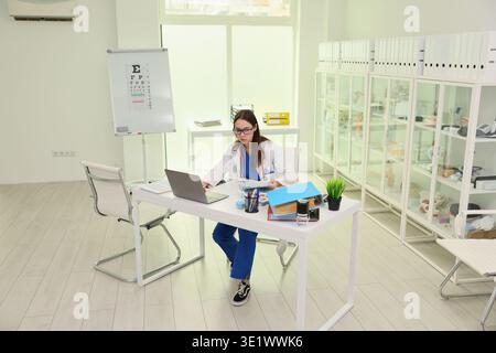 Jeune optométriste féminine travaillant diligemment sur un ordinateur portable dans une clinique oculaire moderne et lumineuse pendant les heures de jour Banque D'Images