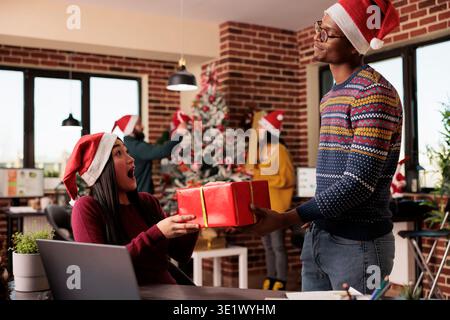 Excité femme asiatique employée dans santa Hat prenant cadeau de Noël d'homme afro-américain collègue tout en travaillant sur ordinateur portable. Divers collègues échangeant des cadeaux au bureau à la veille de noël Banque D'Images