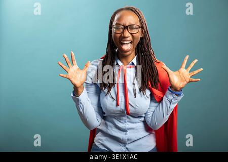 Une dame confiante et puissante pose énergiquement, souriante et riante dans une cape rouge au-dessus d'une chemise bleue. Femme afro-américaine se tient sur fond isolé, exsudant le succès et l'optimisme. Banque D'Images