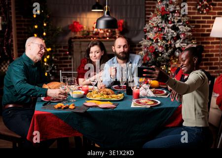 Amis prenant des photos la veille de noël, créant de beaux souvenirs de fête à la maison. Diverses personnes s'amusant avec le vin et la nourriture, appréciant des photos pour des souvenirs à table. Banque D'Images