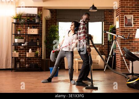 Joyeux couple aime les tâches ménagères que l'homme danse avec l'aspirateur et la femme avec des écouteurs prétend que la vadrouille est une guitare. Heureux partenaires romantiques agissent comme des rockstars tout en nettoyant le salon confortable Banque D'Images
