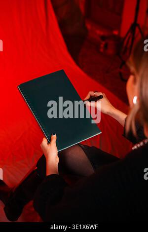 Un fond rouge encadre un cahier vert foncé. Femme le tient proche, prête à écrire. Elle porte du noir, s'assoit bas, les jambes croisées. Son expression suggère Banque D'Images