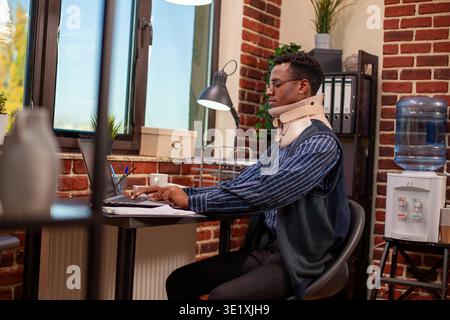 Entrepreneur portant une attelle de cou travaille sur son ordinateur portable dans le bureau de démarrage, se rétablissant d'une blessure et se concentrant sur son projet d'entreprise. Homme d'affaires noir se concentrant sur la recherche d'entreprise après un accident. Banque D'Images