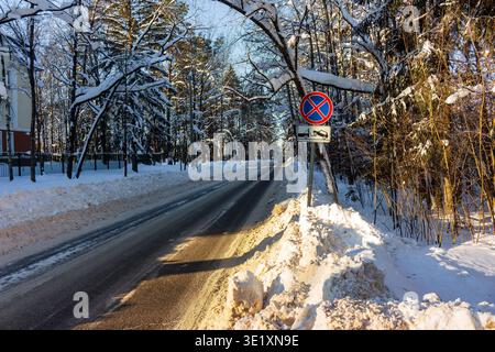 Route d'hiver sereine à travers des arbres chargés de neige. Dégagez le panneau d'interdiction d'arrêt lorsque les supports d'avertissement de remorquage sont bien en vue. Neige embrassée par le soleil et branche givrée Banque D'Images