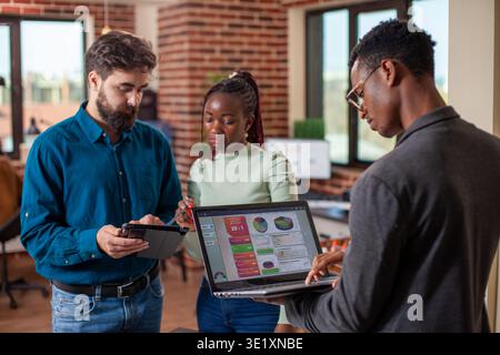 Collègues utilisant la technologie numérique lors de leur discussion dans un espace de travail moderne. Homme noir utilisant un ordinateur portable pour récupérer des données marketing tandis que ses collègues hommes et femmes examinent les plans de stratégie sur tablette. Banque D'Images