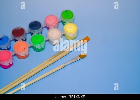 Un ensemble de conteneurs de peinture acrylique ou gouache vibrants avec deux pinceaux en bois sur un fond bleu Uni. Fournitures artistiques créatives pour la peinture pour enfants, les projets scolaires ou les activités de loisirs. Vue de dessus avec espace de copie. De peintures colorées Gouache et pinceaux sur fond bleu. Photo de haute qualité Banque D'Images