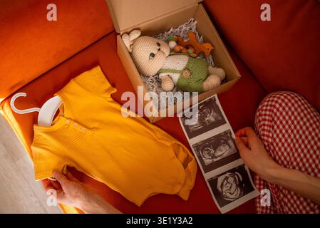 Attendre des parents vus d'en haut tenant une photo échographique et une petite onesie à côté d'une boîte cadeau, se préparant pour leur nouveau-né avec amour, espoir et anticipation confortable à la maison Banque D'Images
