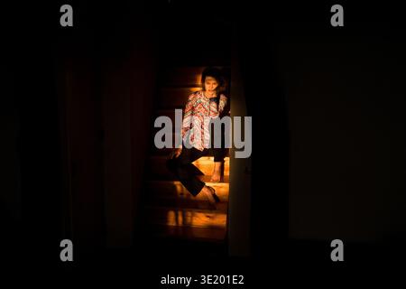 Portrait de mode de vie de la mode d'une jeune femme assise sur des escaliers en bois, éclairée par une lumière douce et chaude qui coule d'une fenêtre voisine alors qu'il fait sombre autour. Banque D'Images