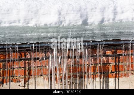 Des poignards gelés de glace coulent au-dessus d'un mur de briques rustique, baignés par un soleil d'hiver éclatant. Danger de fonte de la neige et de la glace Banque D'Images