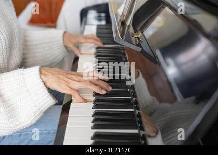Gros plan des mains d'une femme d'âge moyen, portant un pull blanc, jouant d'un piano droit noir et appuyant sur les touches ivoire Banque D'Images