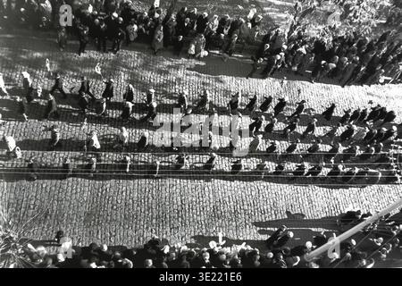 Photographie d'archive dynamique en noir et blanc prise en 1967, représentant une vue en haut-angle de citoyens marchant en rangées nettes lors de la manifestation du 7 novembre dans le centre-ville de Slavyansk. Les participants marchent le long d'une rue pavée historique, projetant de longues ombres clairement définies dans la lumière du soleil d'automne, avec des spectateurs le long de la route. Cette image documentaire captivante préserve la mémoire des événements publics organisés et l’atmosphère nostalgique d’un Donbass pacifique avant la guerre. Banque D'Images