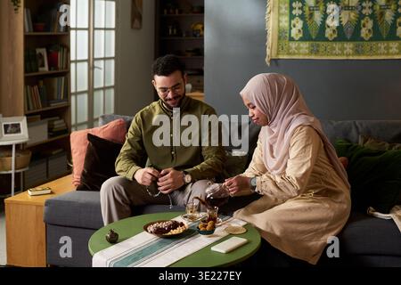 Jeune homme musulman assis sur un canapé souriant tandis que jeune femme musulmane adulte en hijab versant du thé dans une tasse, des collations traditionnelles et une théière sur la table entre eux Banque D'Images