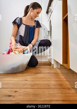 Femme agenouillée, chargement de vêtements dans la machine à laver dans la buanderie moderne. Allemagne Banque D'Images