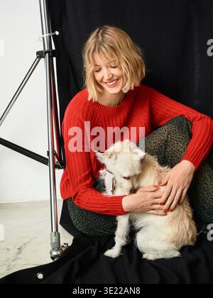 Femme dans un pull rouge câlin un petit chien sur un ensemble de photoshoot avec un fond noir. Allemagne Banque D'Images