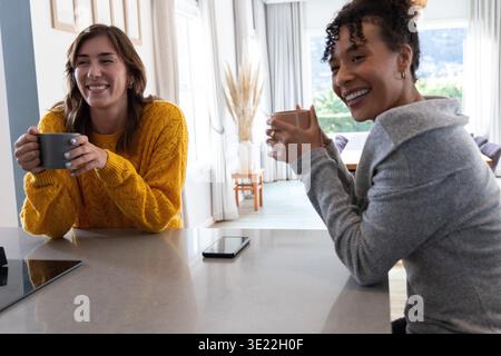Diverses amies femmes sirotant des tasses et discutant à l'îlot de cuisine, smartphone entre eux. Comptoir, femmes, ensoleillé, salle à manger, maison, chaleur, connexion || autorisation du modèle Banque D'Images