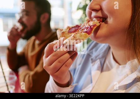 Jeune femme mangeant Pizza Slice en étant assise avec Young Man à l'intérieur Banque D'Images