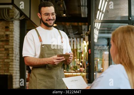 Jeune homme adulte souriant tout en prenant la commande d'une cliente Banque D'Images