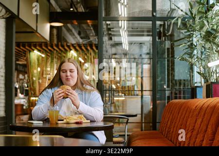 Jeune femme appréciant manger hamburger et frites à table du restaurant Banque D'Images