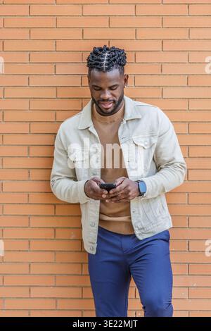 Un jeune homme se tient décontracté appuyé contre un mur de briques, concentré sur son smartphone. Il porte une veste légère sur un pull marron, incarnant une tendance Banque D'Images